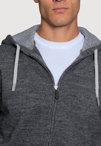 Szara bluza zapinana na zamek z ciemnoszarym podszyciem, widoczna tekstura materiału, sznureczki w jasnoszarym kolorze, noszona na białej koszulce z okrągłym dekoltem.