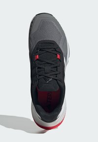 Zapato deportivo gris con parte superior de malla, cordones negros y acentos rojos. Presenta una suela acolchada y punta redondeada.