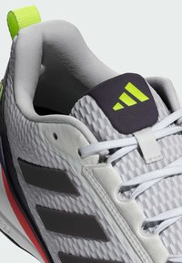 Baskets de sport gris clair avec une tige texturée, des rayures marron foncé, une languette violette et des accents vert fluo. Elles sont dotées de lacets blancs et d'une languette de traction.