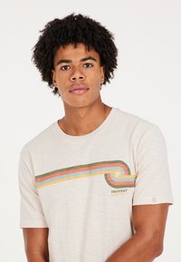 Camiseta de manga corta beige con rayas horizontales multicolores (verde, rosa, azul) y logo "PROTEST". Tejido suave, ajuste casual.