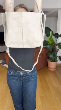 Világosszürke velúr táska, amelyet felemeltek, lapos formájú, hosszú vállpánttal rendelkezik, és finom varrási részletek láthatók egy sima háttér előtt.