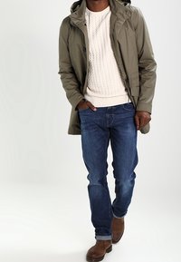 Homme portant une veste à capuche vert olive, un pull en maille torsadée crème, un jean bleu et des bottes en cuir marron, marchant vers l'avant avec une main dans la poche.