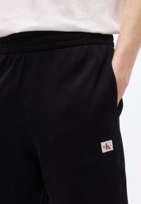 Άτομο που φοράει μαύρο παντελόνι φόρμας με ελαστική μέση και το λογότυπο της Calvin Klein ως ταμπέλα στην πλαϊνή τσέπη, με το χέρι εν μέρει στην τσέπη.