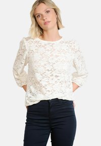 Lace blouse in white with long, puffed sleeves and a round neckline; features floral patterns and a sheer texture. Paired with dark jeans.
