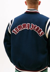 Giubbotto bomber blu navy con strisce bianche e bordeaux. Caratterizzato da una grande scritta ricamata "TOMMY JEANS" sul retro. Tessuto morbido e strutturato.