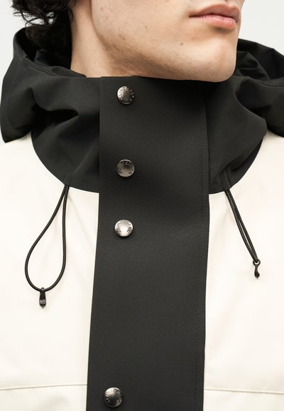 Κοντινό πλάνο ενός ατόμου που φοράει ένα ασπρόμαυρο μπουφάν με ψηλό γιακά, το οποίο ασφαλίζεται με τέσσερα μεταλλικά κουμπιά snap και μαύρα κορδόνια.