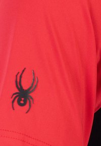 Camisa de manga corta de tela roja con un logotipo de araña negra en la manga izquierda. El material tiene un acabado liso y texturizado.