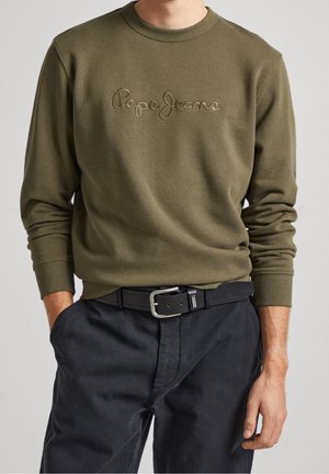 Mężczyzna ubrany w oliwkową bluzę Pepe Jeans i czarne spodnie z czarnym paskiem, stojący z jedną ręką w kieszeni na jednolitym tle.