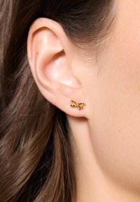 Złote kolczyki, zaprojektowane w kształcie małej kokardy, z misternymi detalami i wypolerowanym wykończeniem, podkreślają urodę ucha.