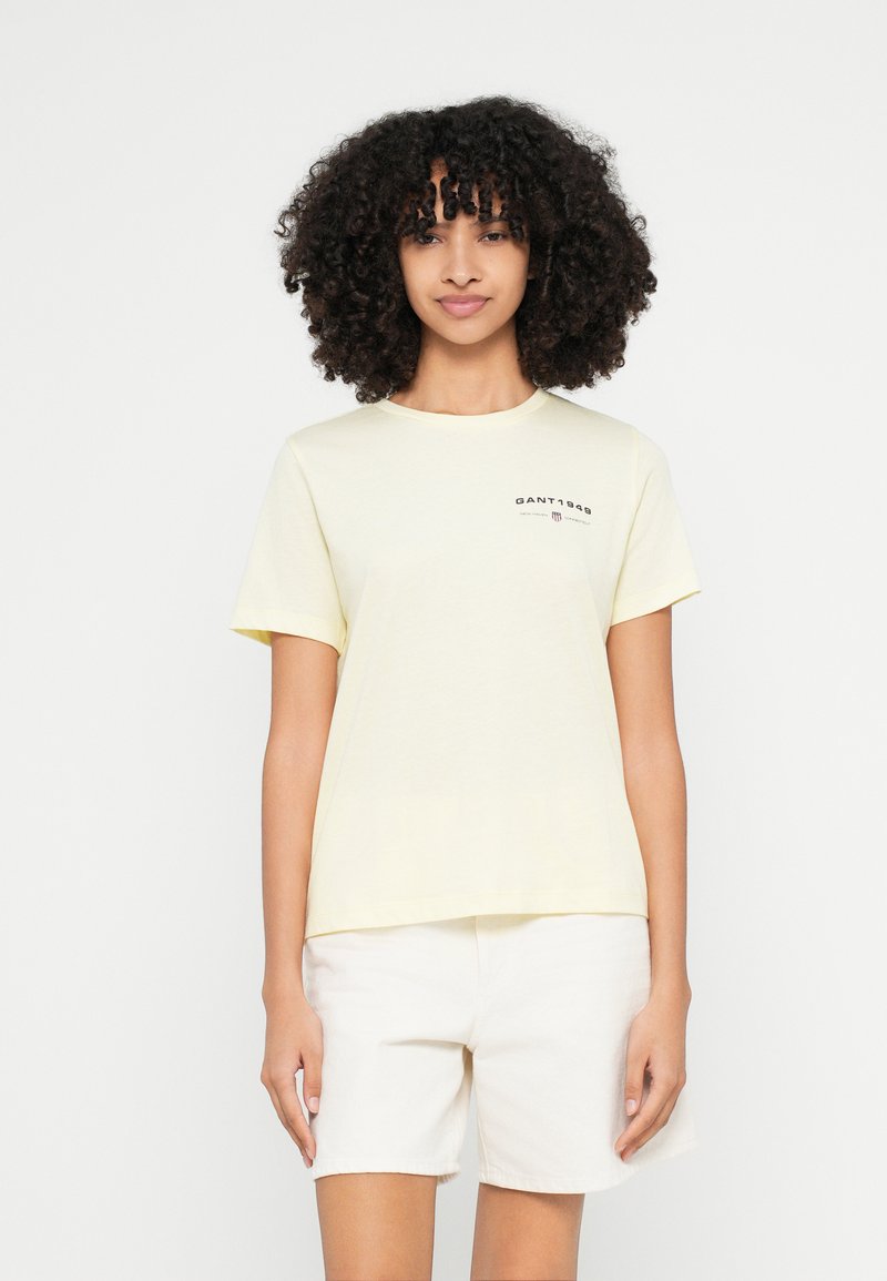 Hellgelbes Baumwoll-T-Shirt mit rundem Halsausschnitt und kurzen Ärmeln, das ein kleines schwarzes Logo auf der Brust trägt. Kombiniert mit weißen Shorts.