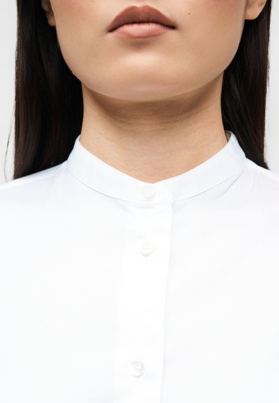 Λευκό πουκάμισο με γιακά stand-up, διακριτή υφή και λεπτομέρειες με κουμπιά κατά μήκος της πατιλέτας. Κοντινή εικόνα του λαιμού και του υφάσματος.