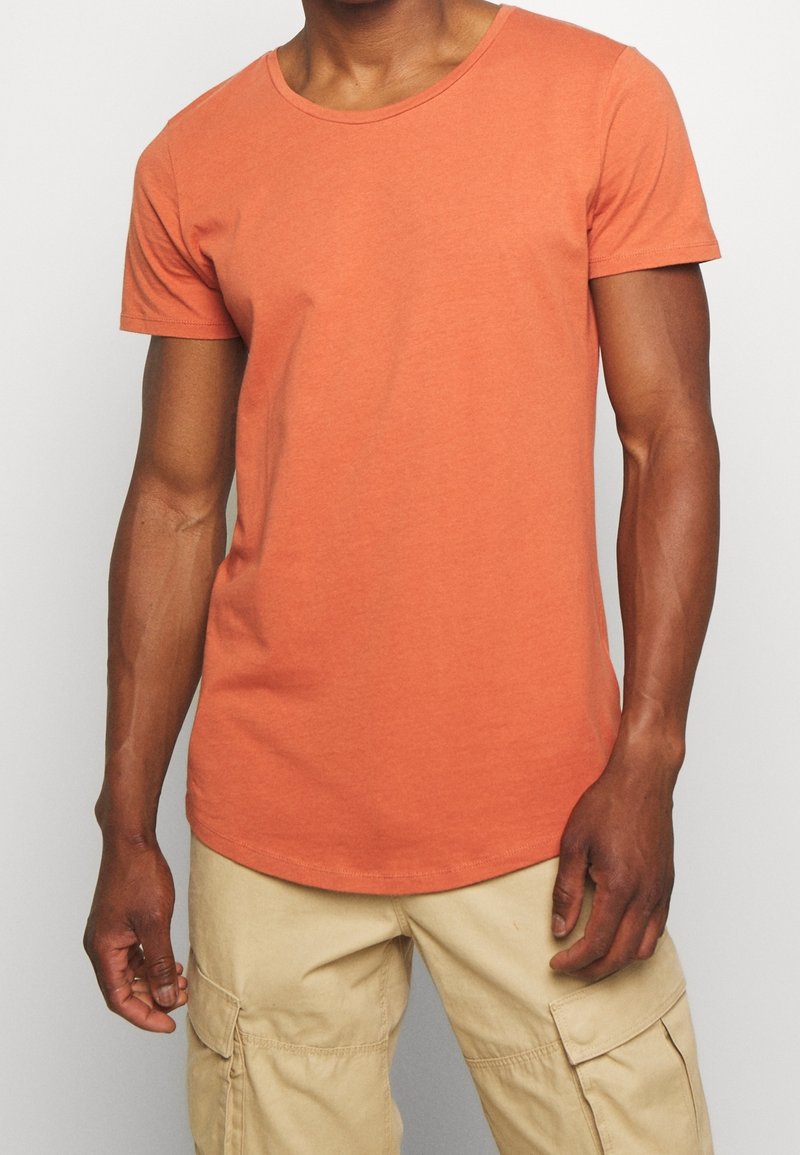 T-shirt à manches courtes de couleur orange clair, en coton doux. Il présente un col rond et une coupe ajustée, porté avec un short cargo beige.
