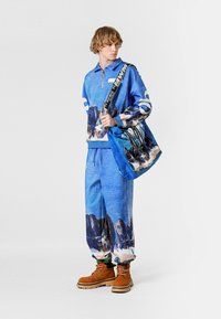 Sudadera azul con cremallera y estampado de montaña, pantalones deportivos azules a juego y botas marrones. La sudadera tiene detalles blancos y un bolso grande de hombro.