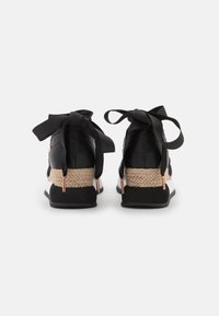 Baskets noires avec des rubans noirs noués en un nœud, dotées d'une semelle intermédiaire beige tissée, d'une empeigne noire texturée et de subtils accents métalliques.