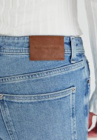 Jeans de mezclilla azul claro con una parche de cuero marrón que tiene el texto en relieve "NOSOTROS LOS LIBRES." Diseño clásico de cinco bolsillos con costuras de contraste.