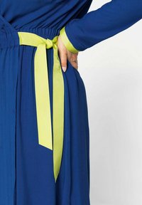 Robe bleu marine en tissu texturé, avec un ruban vert citron noué à la taille et des poignets assortis. Poche latérale visible.