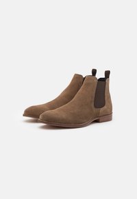 Bottines en daim marron avec des panneaux élastiques sur les côtés, des languettes de tirage et un design à bout rond. Talon en bois plat et détails de couture contrastants.