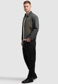 Mladý muž stojaci s rukami vreckách, oblečený v sivej bunde na zips, bielom tričku, čiernych džínsoch a čiernych teniskách na jednoliatom pozadí.