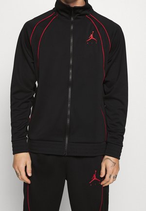 Osoba nosząca czarną rozpinaną bluzę sportową i dopasowane spodnie z czerwonymi lamówkami oraz czerwonymi logo Jumpman Air na lewej piersi i udzie.