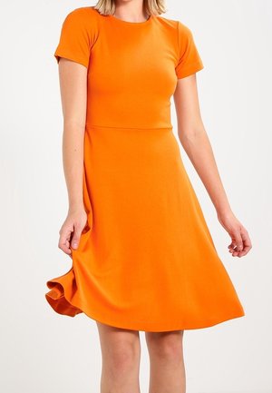 Femme portant une robe orange vif à manches courtes et à hauteur des genoux, tenant l'ourlet tout en se tenant devant un fond blanc uni.