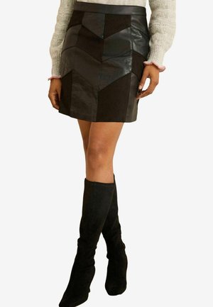 Schwarzer Leder-Minirock mit einem geometrischen Patchwork-Design, das eine Mischung aus matten und strukturierten Oberflächen aufweist. Kombiniert mit schwarzen Overknee-Stiefeln.