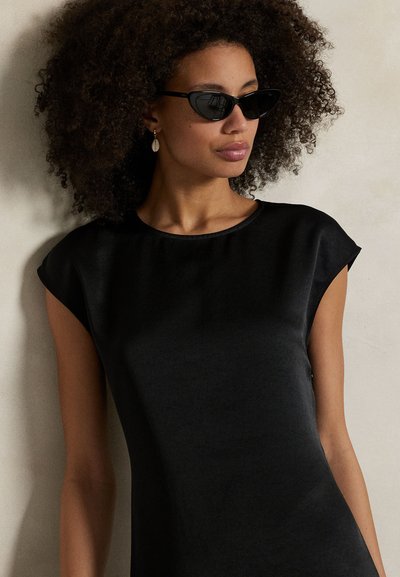 Vestido preto com textura lisa, mangas tipo capa e decote arredondado. Usado com óculos de sol estilo olhos de gato e um brinco pendente.