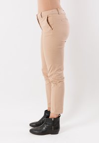 Vista laterale di una persona che indossa pantaloni slim-fit beige e stivaletti in pelle nera con cerniere laterali, su uno sfondo bianco neutro.