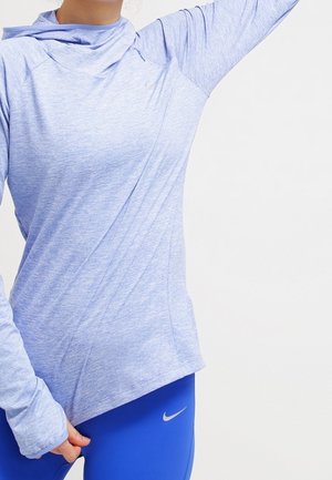 Personne portant un haut de sport bleu clair à manches longues et des leggings Nike bleu vif, levant le bras et tenant l'ourlet du haut.