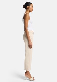 Pantalons beiges taille haute avec une jambe droite et des poches arrière discrètes, associés à un débardeur blanc à côtes et des chaussures à talons beiges.
