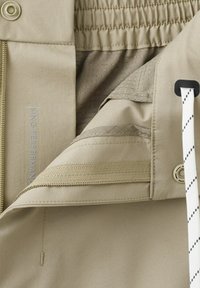 Primer plano de la cintura de unos pantalones beige con detalles elásticos, botón, cremallera y cordón blanco con rayas diagonales negras.