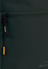 Donkergroene rugzak van textuurstof, met een ritsvak, zwarte trekhaak en een oranje accent bij de rits. Logo aan de onderkant gedrukt.