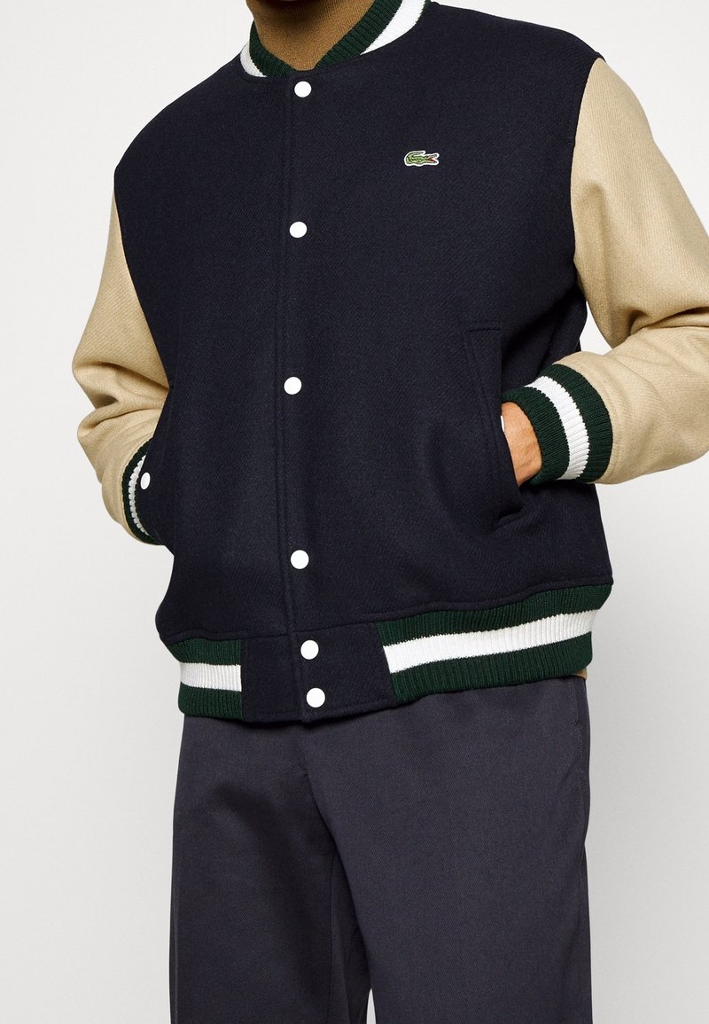 Homme portant une veste varsity bleu marine et beige avec des boutons-pression blancs, des poignets et un ourlet rayés, et un petit logo crocodile vert sur la poitrine.