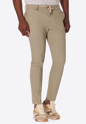 Uomo che indossa pantaloni beige slim fit con coulisse in vita e sneakers beige e bianche, in piedi su uno sfondo bianco.