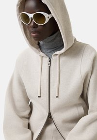 Beige lynlås-hoodie med hætte, der har en grå rullekravebluse underneath, suppleret af hvide ovale solbriller med tonede linser.