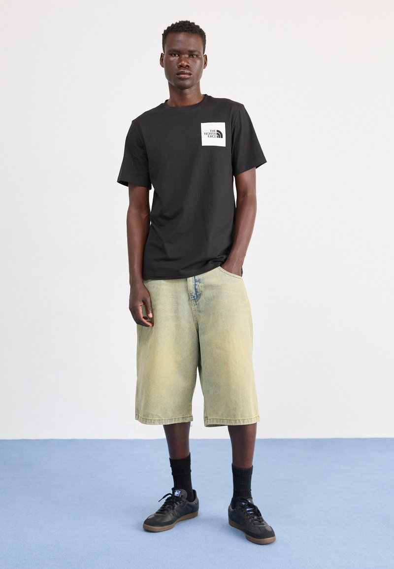 Czarna bawełniana koszulka z białym logo na piersi, w połączeniu z jasnymi żółtymi szortami dżinsowymi i czarnymi trampkami. Jednolity kolor, swobodny krój.