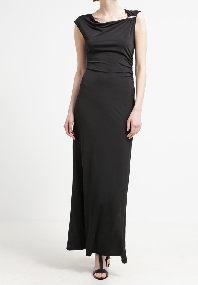 Femme portant une robe noire longue sans manches, avec un décolleté drapé et des côtés froncés, associée à des sandales noires à lanières.