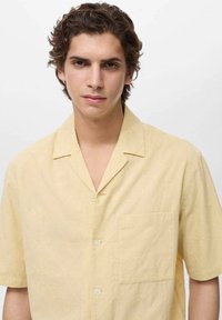 Korte mouwen, lichtgele shirt met een zak en een subtiele textuur met bloemmotief. Knopsluiting aan de voorkant en een camp collar ontwerp.