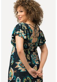 Fließendes Kleid mit dunklem Hintergrund, der bunte geometrische Muster zeigt. Auffälliges Schnürdetail im Rücken und Rüschensäume.