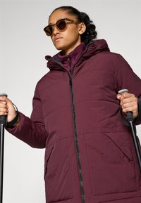 Doudoune bordeaux avec fermeture éclair à l'avant, capuche et poches ; le modèle tient des bâtons de trekking, porte des lunettes de soleil marron. Le matériau semble doux et texturé.