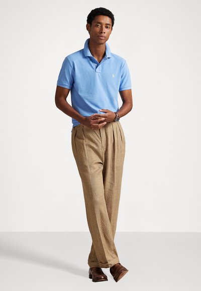 Ανοιχτό μπλε πόλο μπλούζα με γιακά, συνδυασμένη με μπεζ καρό παντελόνι. Το σύνολο έχει χαλαρή εφαρμογή και καφέ δερμάτινα παπούτσια.