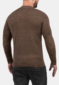 Brun strikket sweater med lange ærmer, ribbede manchetter og kant, albuepatcher og et lille logo-tag nederst på ryggen. Glat tekstur.