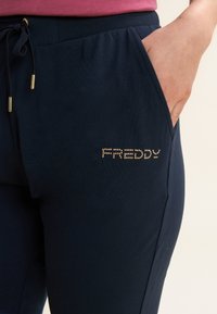 Pantalones deportivos azul marino hechos de tela suave, con cintura elástica, cordón ajustable, bolsillos laterales y logotipo "FREDDY" con tachuelas doradas.