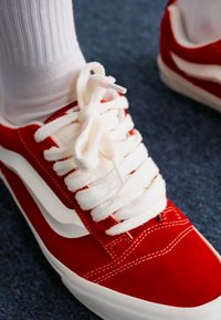 Piros velúr sportcipő fehér bőr részletekkel, vastag fehér fűzőkkel és látható varrással. Texturált felület, kék háttéren.