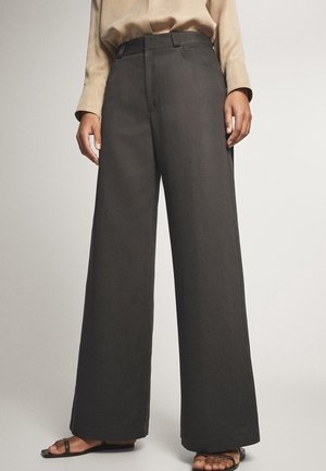 Personne portant un pantalon gris foncé taille haute à jambes larges, associé à un haut à manches longues beige et des sandales noires à bouts ouverts.