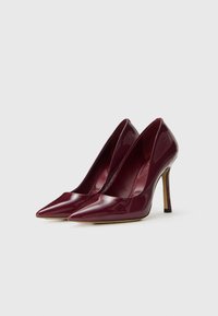 Une paire de chaussures à talons aiguilles brillantes de couleur rouge foncé, avec des talons stilettos, présentées côte à côte sur un fond blanc.