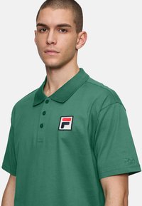 Grön pikétröja i bomull med knäppning, med broderad Fila-logga på bröstet och text på ärmen. Ren design.