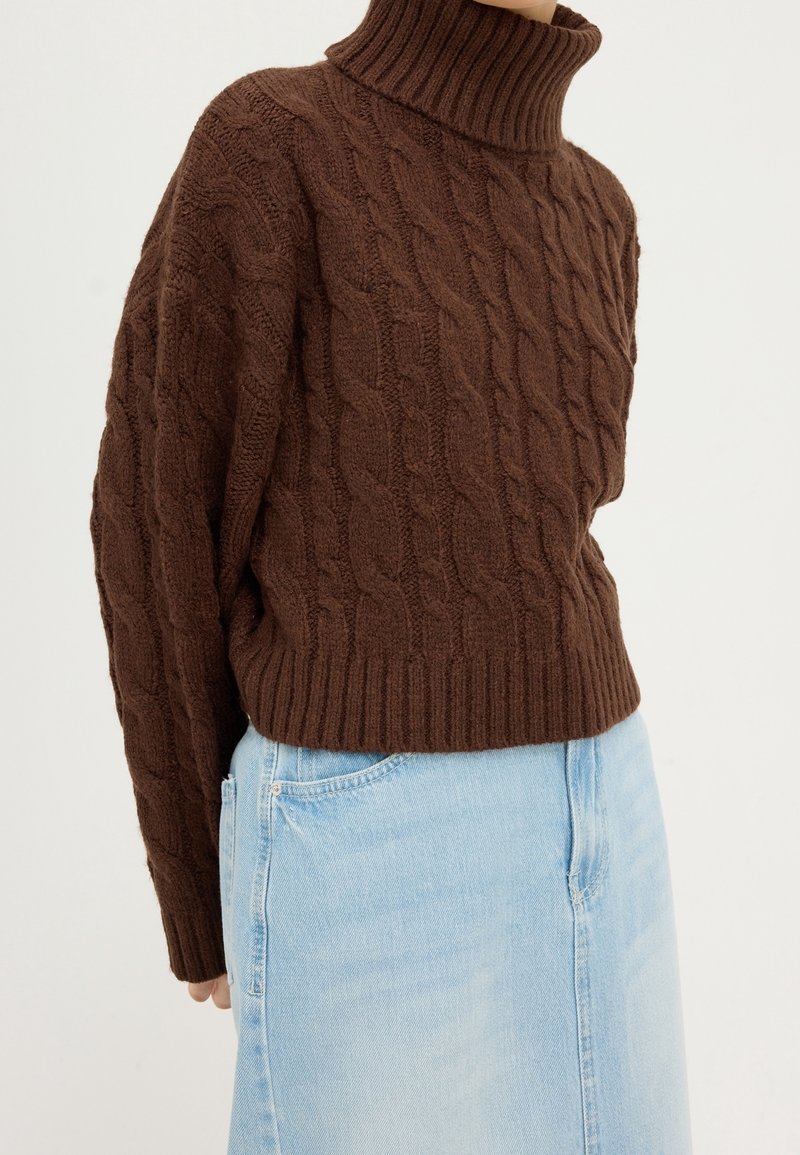 Hnědý svetr s pleteným vzorem a vysokým žebrovaným límcem, s texturovanými vzory a volným střihem, nošený přes světle modrou džínsovou sukni.