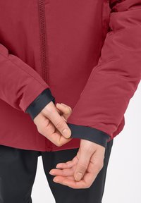 Veste isolante rouge au toucher lisse avec fermeture éclair. Les poignets élastiques noirs sont visibles sur les manches, soulignant un design ajusté.