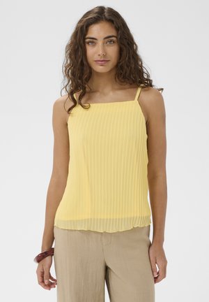 Femme aux longs cheveux bouclés portant un haut plissé sans manches jaune et un pantalon beige, debout devant un fond clair uni.