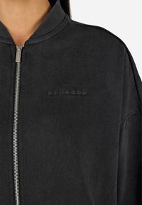 Čierna bunda na zips s ryhľovaným golierom, vyrobená z mäkkej tkaniny. Má textúrované písmená na hrudi a strieborné zipsové kovanie.